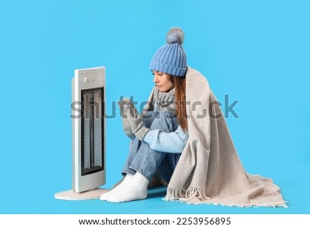 Frozen young woman warming near electric heater on blue background Royalty-Free Stock Photo #2253956895