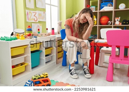 Young blonde woman teacher sitting on chair with stressed expression at kindergarten