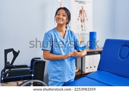 Young chinese woman wearing physiotherapist uniform using touchpad at rehab clinic