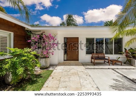 Beautiful entrance of house with cement and stone floors, tropical plants, windows, outdoor chair and table, white walls, blue sky located in Pinecrest, Miami-Dade Royalty-Free Stock Photo #2253993435