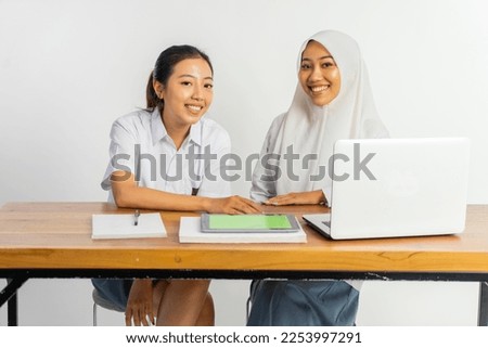 two high school girls sitting at desk with laptop, tablet and books on isolated background Royalty-Free Stock Photo #2253997291