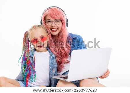 Photo of delighted mother in glasses and pink wig and headphones looks at camera holding laptop on knees sitting on white floor in studio with preschooler daughter with colorful braids and sunglasses.