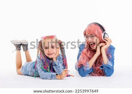 Young modern mother and preschooler daughter in good mood laying on white copy space floor in studio watching movie cartoon. Girls in denim with colorful braids pink wig and sunglasses.