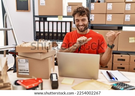 Young hispanic call center agent man working at warehouse pointing to the back behind with hand and thumbs up, smiling confident 