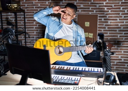 Hispanic young man playing classic guitar at music studio doing peace symbol with fingers over face, smiling cheerful showing victory 