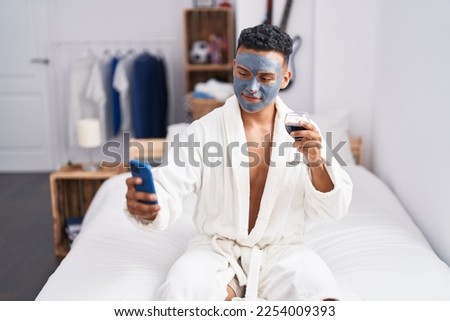 Young latin man sitting on bed make selfie by smartphone drinking wine at bedroom