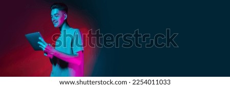 Young smiling boy in casual T-shirt looking on tablet, working, studying remotely over gradient background in neon light. Concept of emotions, occupation and youth lifestyle. Banner, flyer, ad