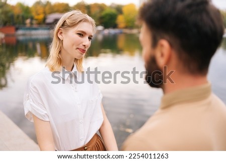 Close up view from back of unrecognizable bearded man to cheerful woman standing holding hands with boyfriend outdoors by beautiful lake at city park, talking and looking at each other smiling.