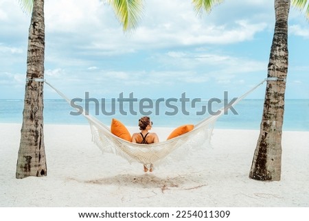 Young woman relaxing in wicker hammock on the sandy beach on Mauritius coast and enjoying wide ocean view waves. Exotic countries vacation and mental health concept image. Royalty-Free Stock Photo #2254011309