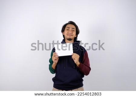 Souteast asian man wearing a sweater and holding blank sheet of paper. Pointing at paper sheet and looking at camera on white background. For promotion concept. Isolated.