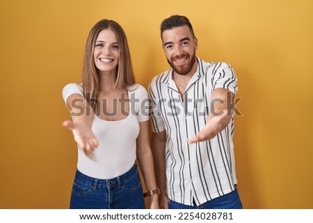 Young couple standing over yellow background smiling cheerful offering palm hand giving assistance and acceptance. 