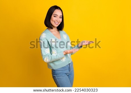 Photo of chinese youngster promoter girl wear stylish daisy print jumper hold palms show empty space new product isolated on yellow color background