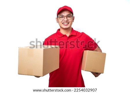 Smiling asian courier man wearing red uniform standing over white background holding two boxes with orders, packing shipping items for clients. Royalty-Free Stock Photo #2254032907