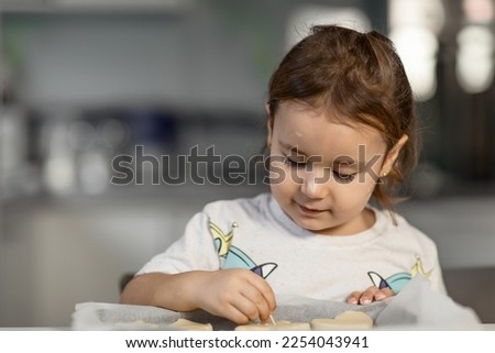 A cute little girl uses a stick to draw drawings on a shortbread dough cut out with metal molds for making homemade cookies