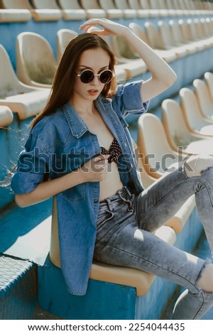 a teenage girl with long dark hair poses for the camera; sits in a spectator seat at the stadium; a model-looking girl on the street; slender figure of a beautiful girl in a bikini under denim clothes