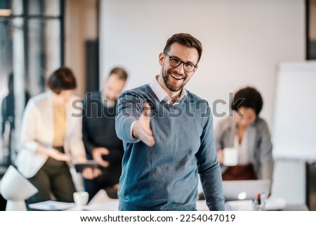 A happy businessman introducing himself while standing at the office with coworkers in blurry background. Royalty-Free Stock Photo #2254047019