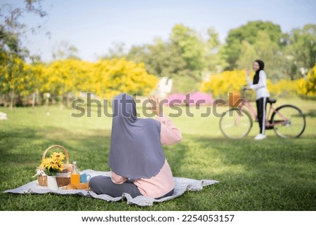 Muslim girl lift her hand to say hi to her friend that riding bicycle far away at park, Islamic friendly, relaxing activity concept and space.