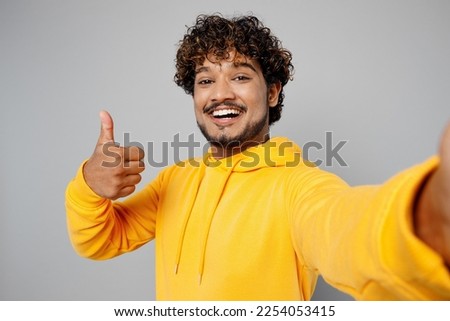 Close up young smiling happy Indian man 20s he wearing casual yellow hoody doing selfie shot pov on mobile cell phone show thumb up isolated on plain grey background studio. People lifestyle portrait