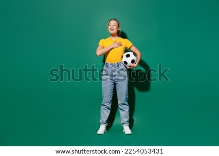 Full body young woman fan wear basic yellow t-shirt cheer up support football sport team hold soccer ball watch tv live stream put hand in heart sing hymn song isolated on dark green background studio Royalty-Free Stock Photo #2254053431