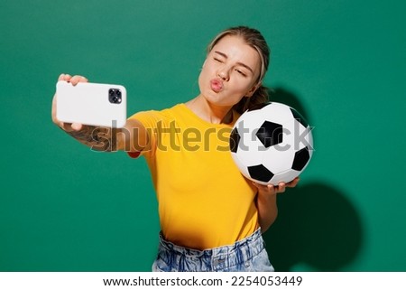Young fun woman fan wear basic yellow t-shirt cheer up support football sport team hold soccer ball watch tv live stream do duckface selfie shot on mobile cell phone isolated on dark green background
