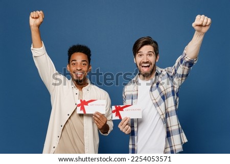 Young two friends excited surprised shocked happy men wear casual shirts together hold gift certificate coupon voucher card for store do winner gesture isolated plain dark royal navy blue background