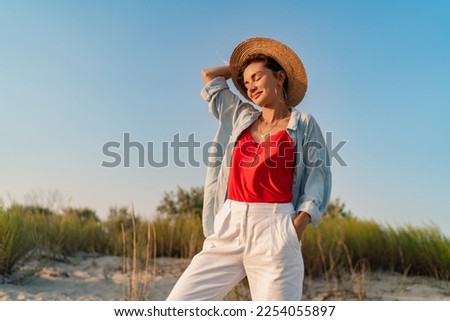 stylish attractive slim smiling woman on beach in summer style fashion trend outfit happy having fun wearing white pants red top blue shirt boho style chic and straw hat