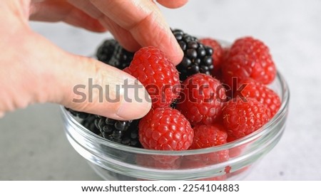 Glass bowl with fresh ripe raspberries and blackberries on a light gray background, woman hands