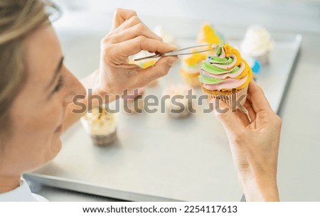 women in pastry bakery as confectioner draped candy pearl topping on a colorful muffin  with tweezers