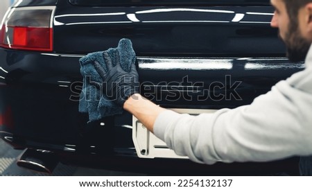 White man wearing black gloves crouching behind black car applying ceramic coating. Professional car detailing process. Horizontal indoor shot. High quality photo