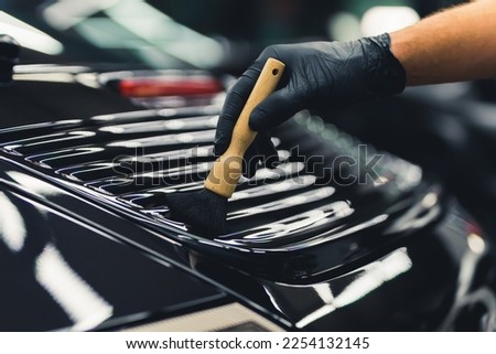 Close-up shot of male hand in black glove wiping brush along detail on trunk of black car. Precision detail work. Car detailing concept. Horizontal indoor shot. High quality photo