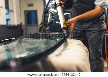 Unrecognisable man applying industrial adhesive to window pane before installing into car. Garage work. Focus on background. Horizontal indoor shot. High quality photo