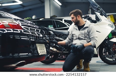 White man kneeling down applying ceramic coating to the back of a black car with motorcycle in the background. Professional car detailing. Garage. Horizontal indoor shot. High quality photo