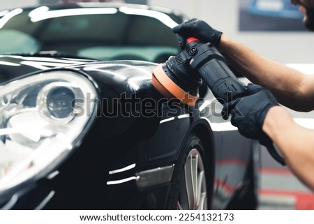 Close-up shot of man wearing black gloves polishing paintwork on the front of a black car using professional tool. Car detailing process. Horizontal indoor shot. High quality photo
