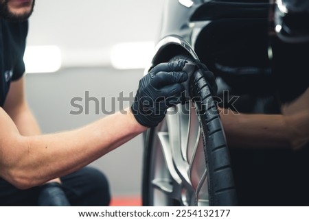 Caucasian man wearing black gloves crouched blackening car tire using sponge. Car detailing in professional garage. Horizontal indoor close-up shot. High quality photo