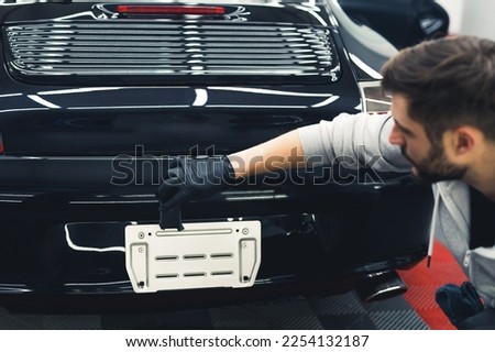 White man wearing black gloves squatting applying ceramic coating to the back of black car. Professional car detailing. Horizontal indoor shot . High quality photo