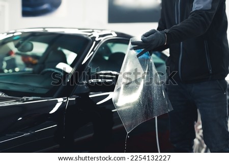 Man wearing black gloves preparing sheet of PPF Paint Protection Film before applying it to side car mirror of black car. Car detailing process. Horizontal indoor shot. High quality photo