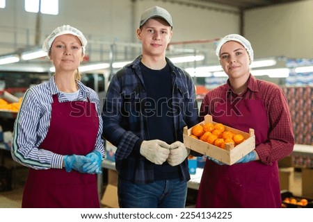 Group of positive workers of fruit sorting and packaging factory standing together in workshop, holding crates with selected ripe tangerines Royalty-Free Stock Photo #2254134225
