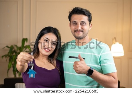Smiling wife with husbnd showing house keys with thumbs up gesture by looking at camera at home - concept of insurance, new home purchase and investment.