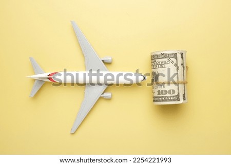 Flatlay picture of toy aeroplane, toy camera, luggage with roll fake money on yellow background. Expensive flight fare.