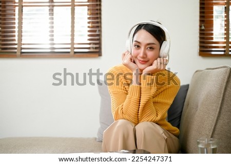 Pretty and charming millennial asian woman in casual clothes relaxing on sofa in her living room, listening to music through her wireless headphones.