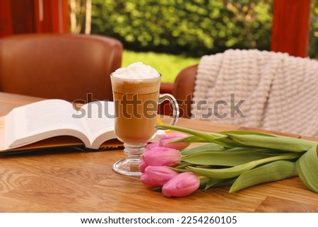 Glass of delicious cocoa, pink tulips and book on wooden table at terrace