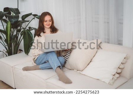 Beautiful cheerful redhead girl using silver laptop while sitting on sofa in living room at home. High quality photo