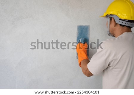 A man wears hard hat and gloves holding trowel is plastering cement wall with copy space for text, concept of wall plastering, structural work, plaster work, home building, construction work  Royalty-Free Stock Photo #2254304101