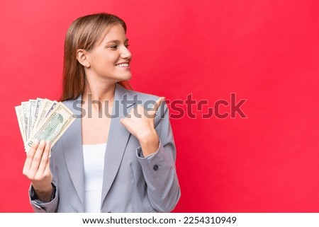 Young business caucasian woman holding money isolated on red background pointing to the side to present a product