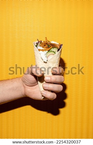 Close-up of a fastfood pita gyros sandwich held by a male hand, yellow background, flash Royalty-Free Stock Photo #2254320037