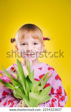 Cheerful little girl in pink sweatshirt with red hearts pattern with bouquet of pink tulips isolated over yellow background. Happy smiling small girl posing in studio.