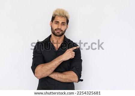 Portrait of positive young man showing direction over white background. Caucasian guy with stubble and highlighted hair wearing black T-shirt pointing finger aside and smiling. Advertising concept