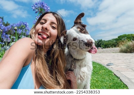 Selfie portrait of young Colombian Latina woman, with her border collie dog, in the park sticking out her tongue and looking at the camera, with the sky and trees in the background, phone perspective. Royalty-Free Stock Photo #2254338353