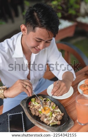 A smiling Filipino man dines in a restaurant and shows how to eat the delicious sizzling pork sisig by mixing the egg with the other ingredients. Royalty-Free Stock Photo #2254342155