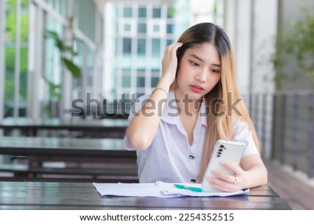 Portrait of cute Asian Thai girl student in uniform is sitting work smiling happily and confidently while using smartphone in the building at university with background.